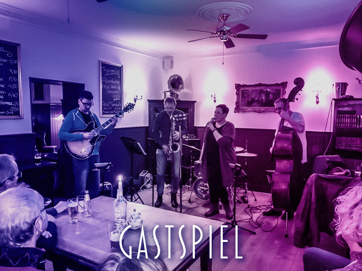 Jazzband mit Gitarre, Saxophon, Gesang und Kontrabass während einer Aufführung in einem Veranstaltungsraum.