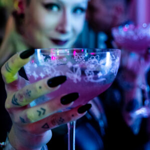 Eine Person hält ein Glas mit pinkem Cocktail, verziert mit floralen Mustern und langen Nägeln.