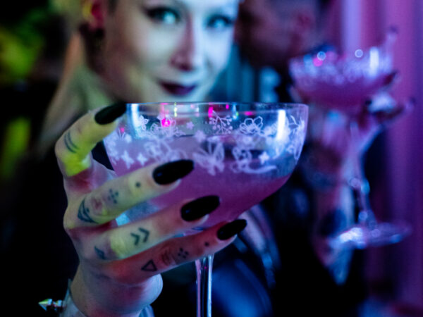 Eine Person hält ein Glas mit pinkem Cocktail, verziert mit floralen Mustern und langen Nägeln.