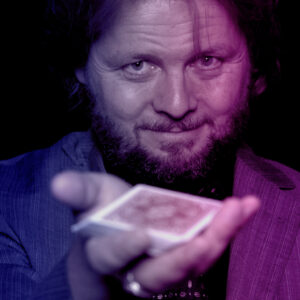 Ein Mann mit Bart zeigt eine Spielkarte in der Hand und lächelt in die Kamera.