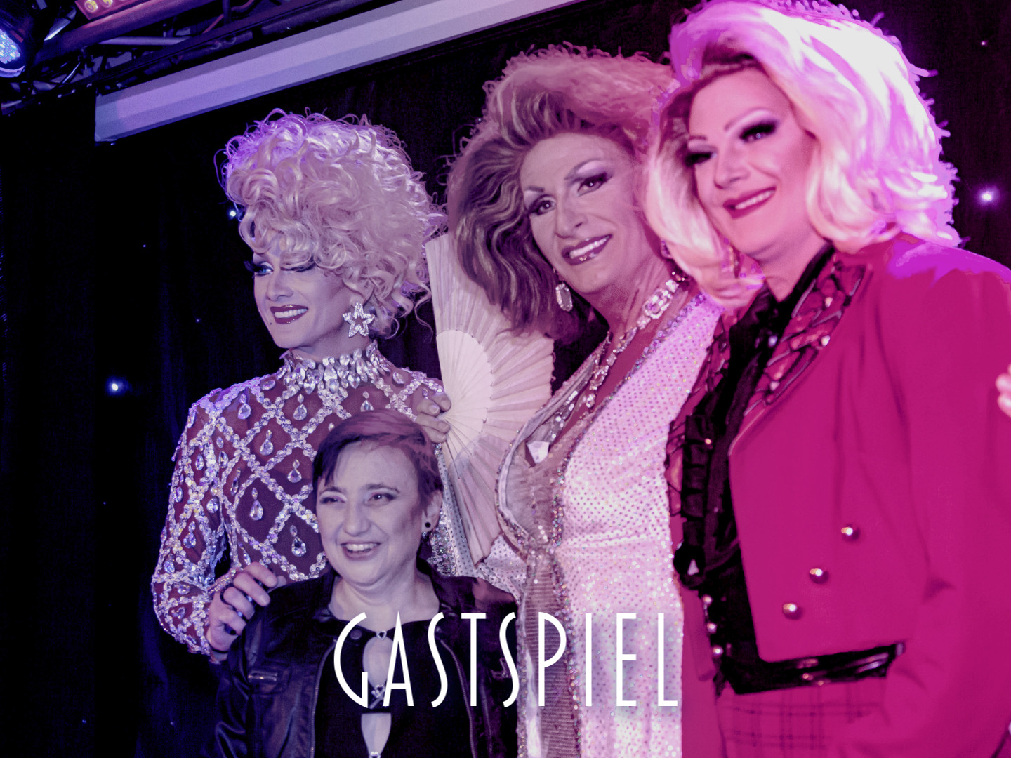 Drei Drag-Künstler in glamourösen Outfits posieren mit einer Person in der Mitte während einer Veranstaltung.