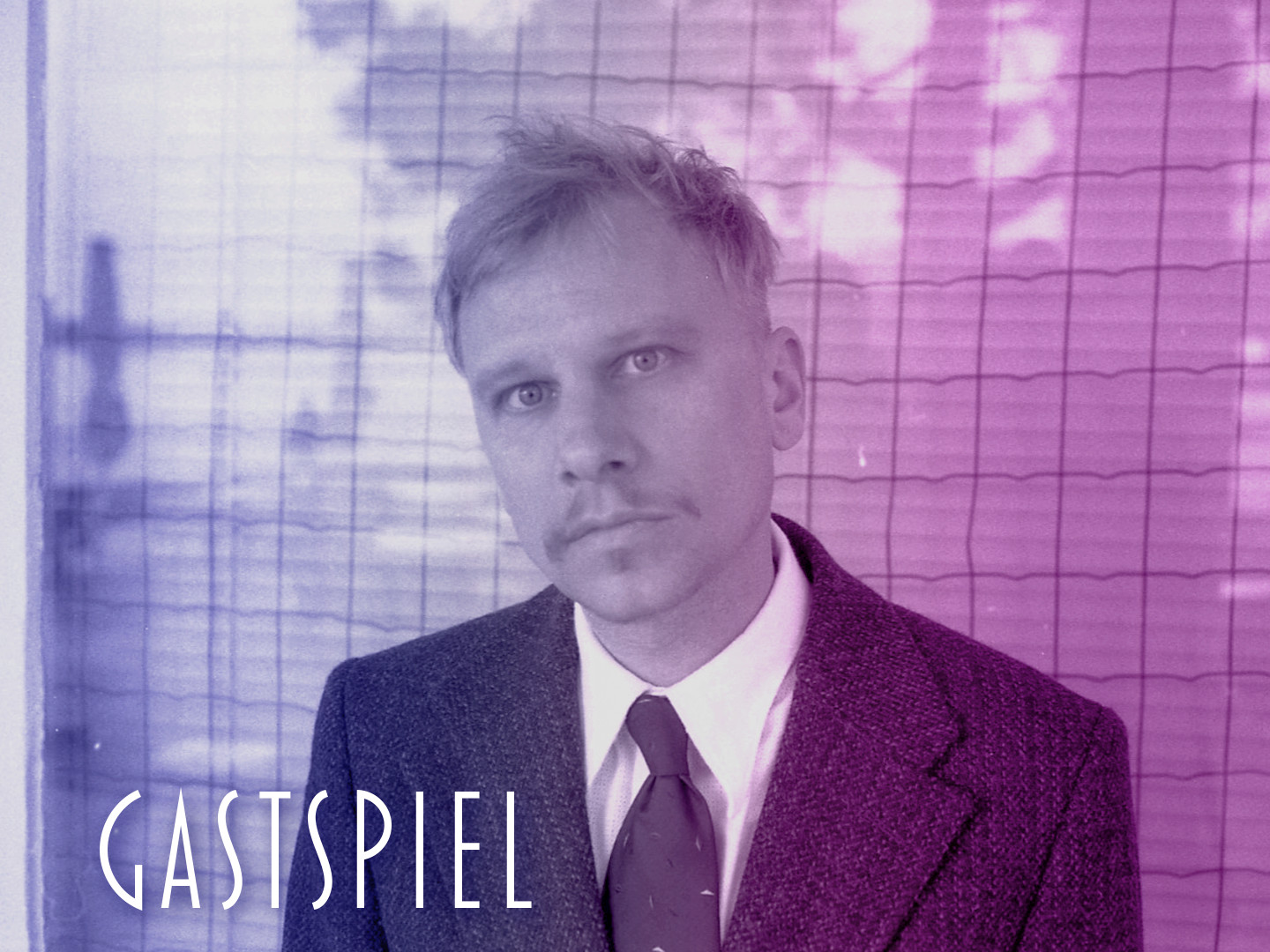 Mann mit blonden Haaren und Anzug, der in die Kamera schaut, vor einem Gitterhintergrund.